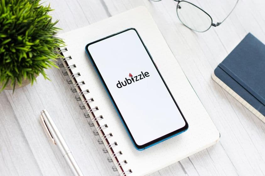 在桌上运行Dubizzle的手机,放在笔记本下,旁边有眼镜和一株小绿植。