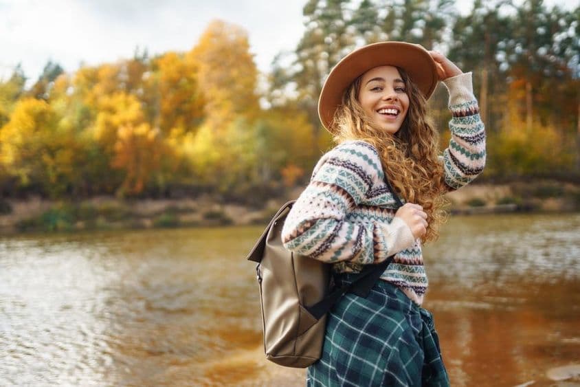 戴帽子和背包的女性旅行者享受秋季沿河徒步旅行。